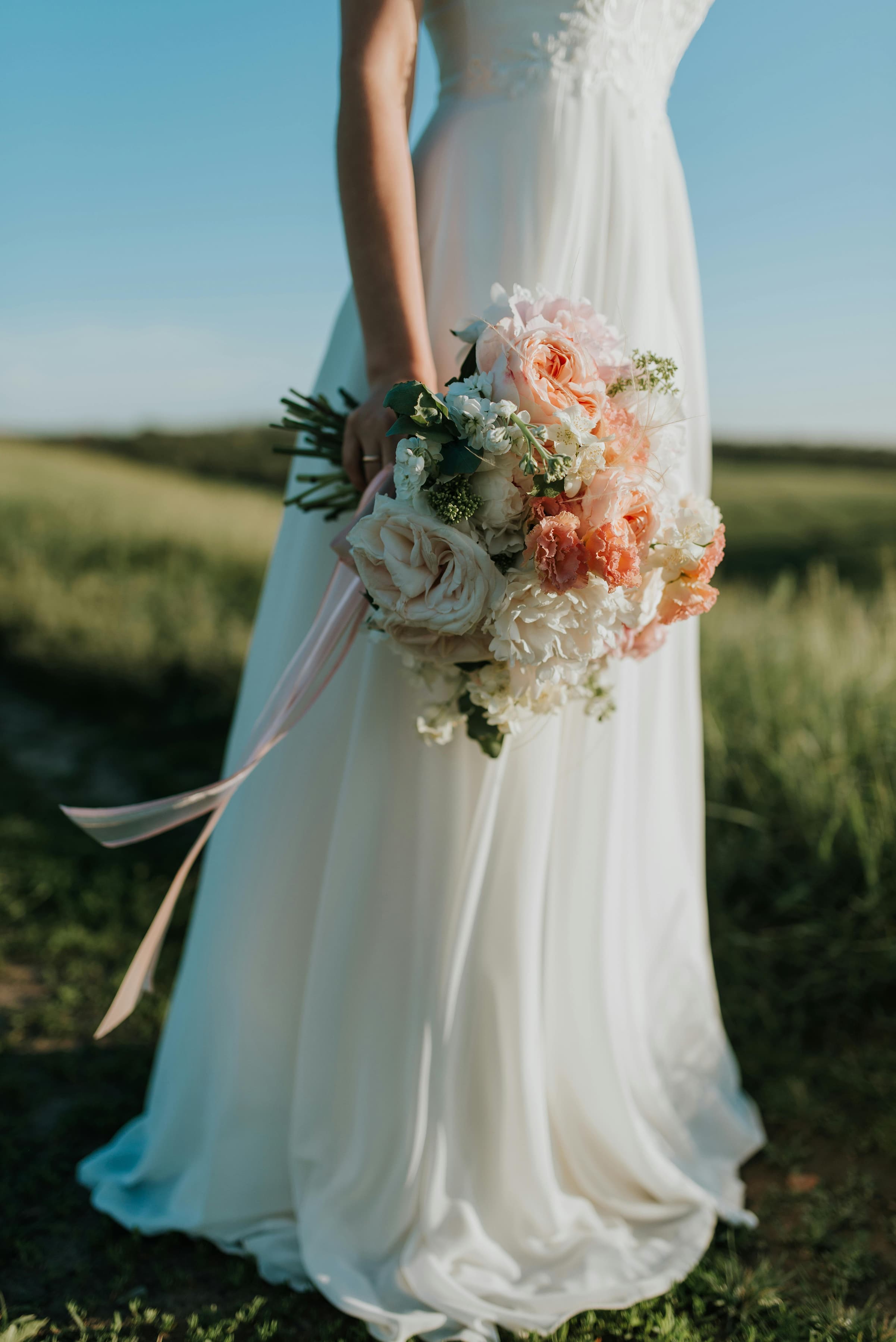 Foto de uma noiva com vestido branco elegante segurando um buquê de flores em um campo ao ar livre.