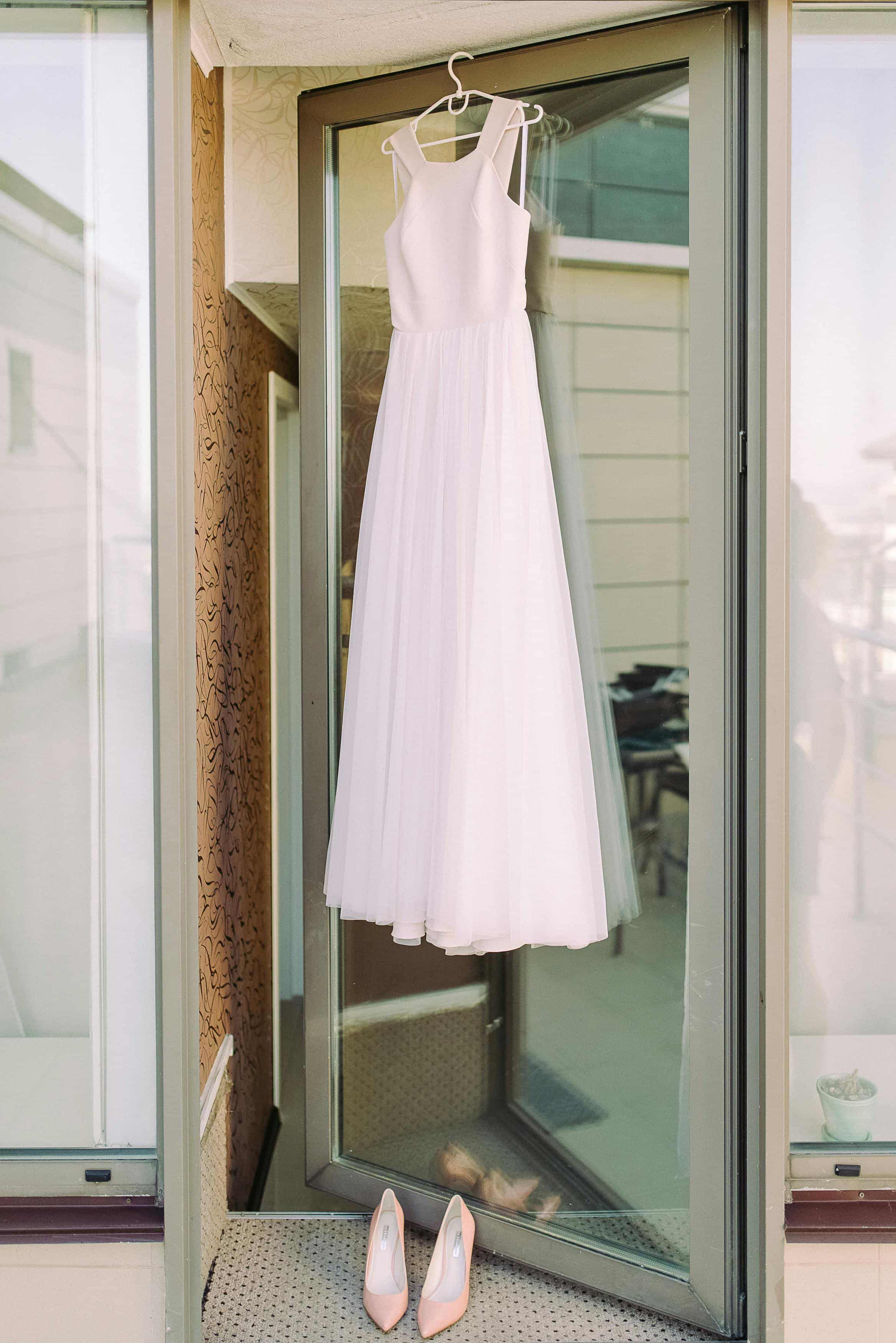 Foto de um vestido de noiva branco delicado e salto alto cor de rosa, pendurados na porta de um closet, pronto para o casamento.