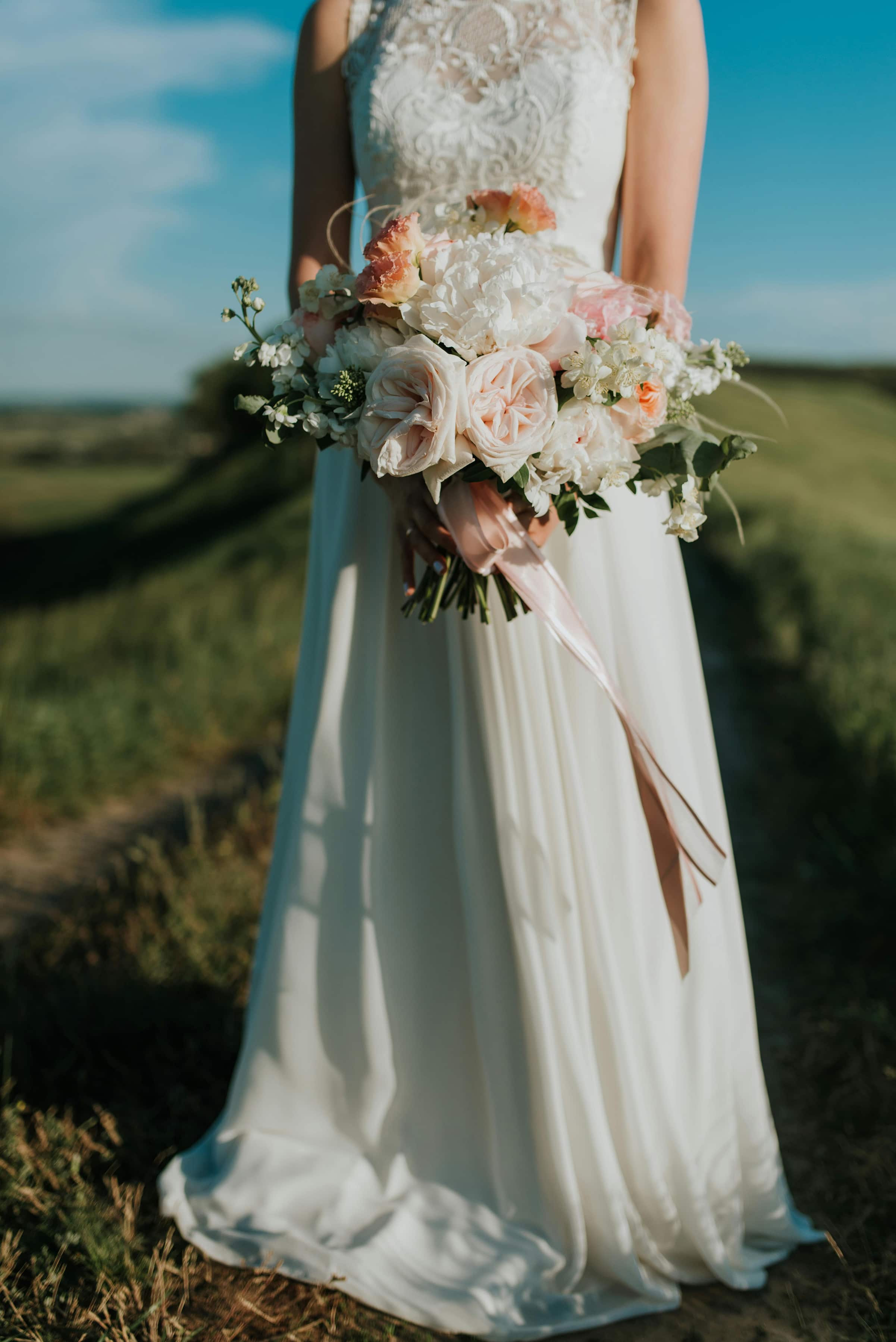 Foto de uma noiva segurando um buquê de flores em um cenário ao ar livre, com vestido de noiva branco e céu azul ao fundo, um casamento ao ar livre.