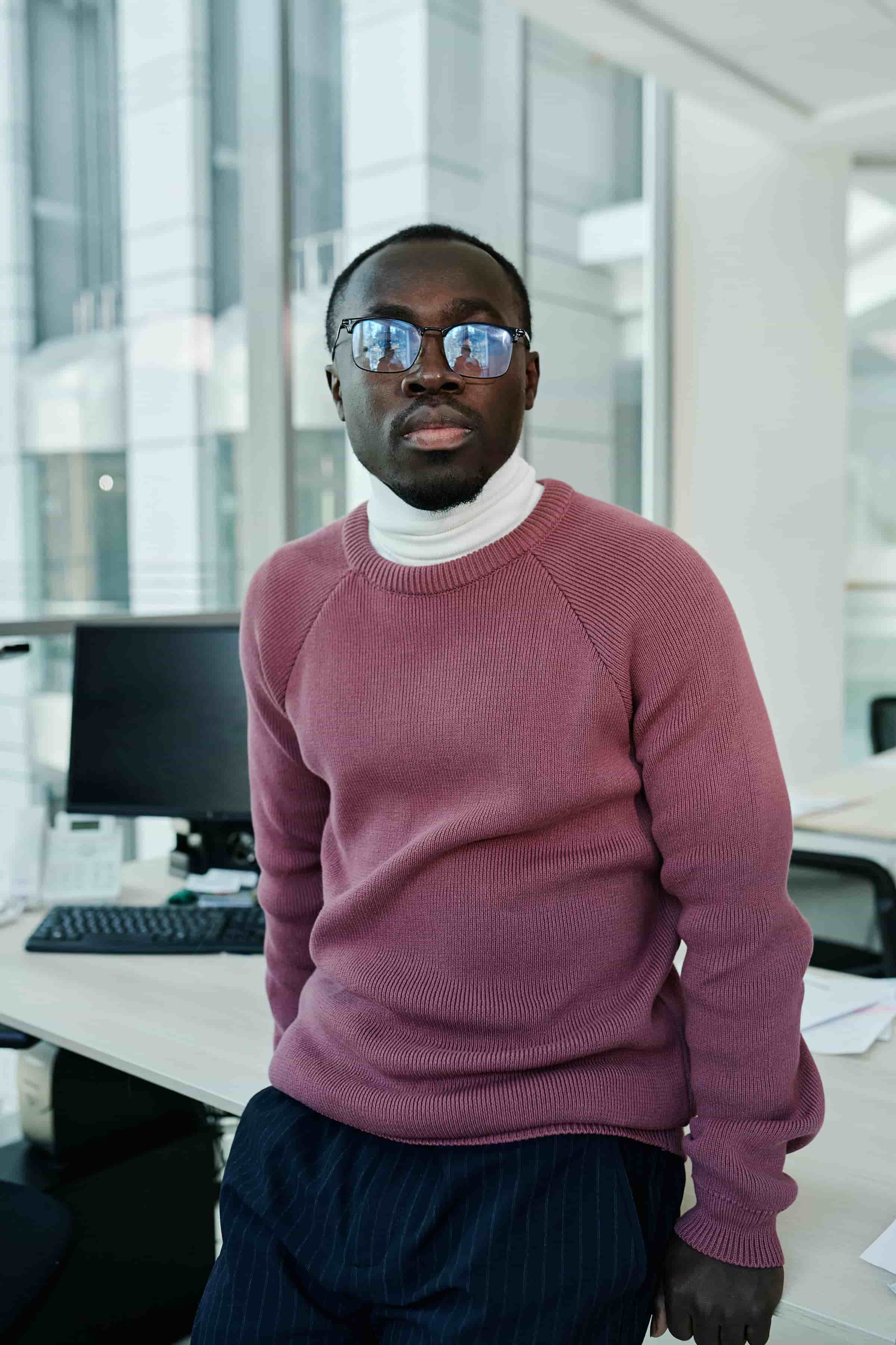 Foto de um homem de óculos escuros e suéter rosa em ambiente de escritório moderno.