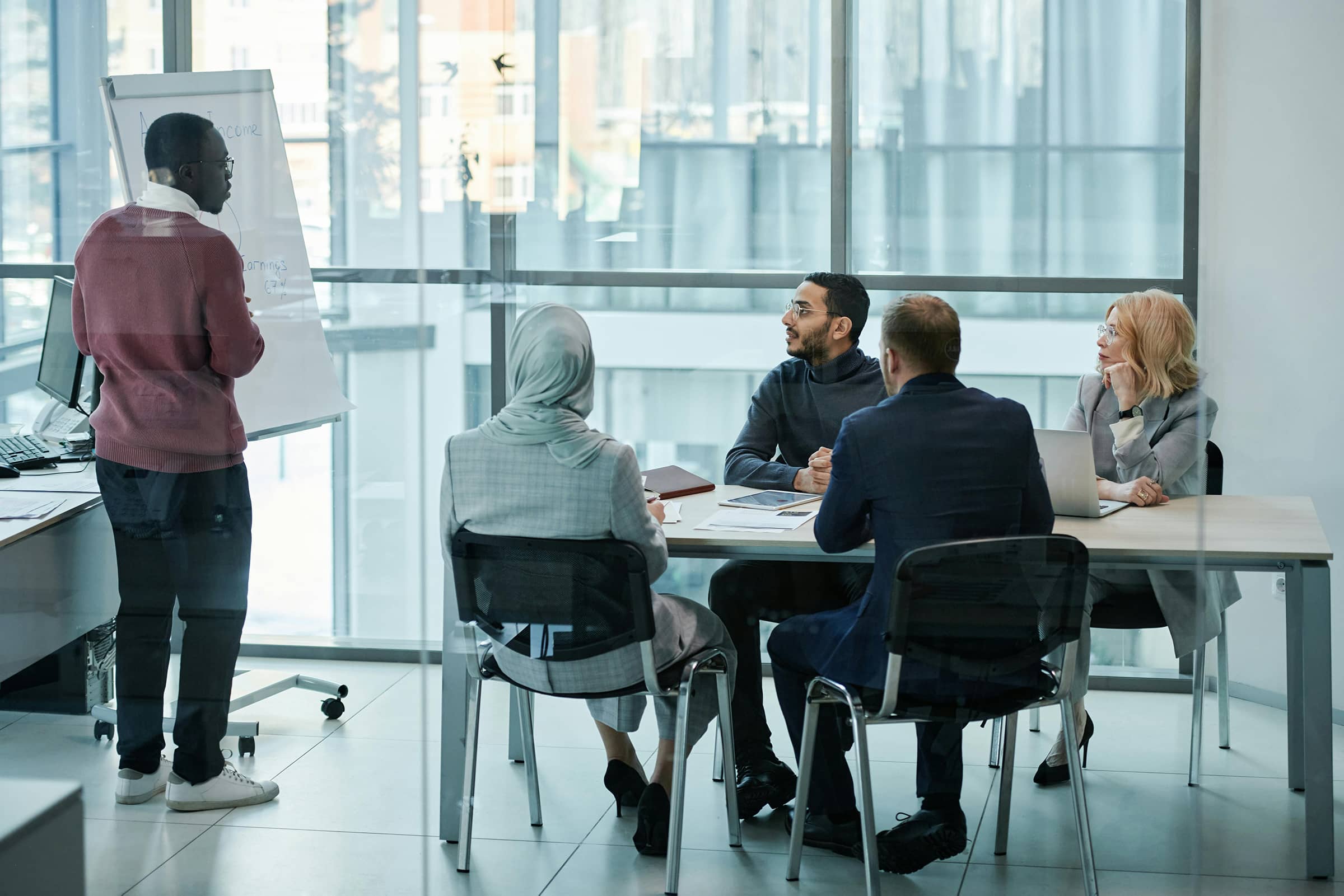 Foto de uma reunião de negócios com diversidade cultural, com uma pessoa apresentando em frente ao grupo em uma sala moderna com vista urbana.