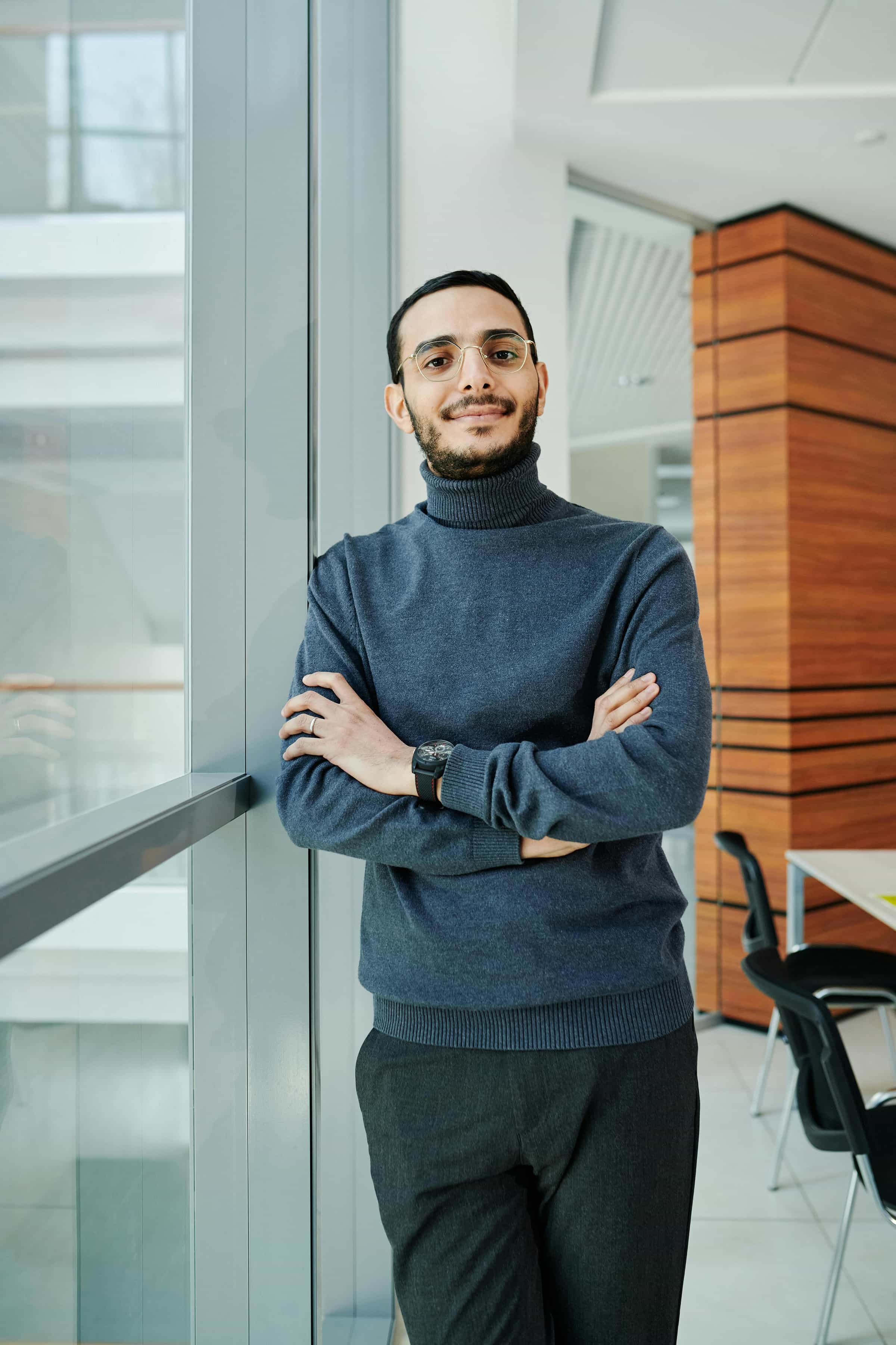 Foto de um homem de camiseta de gola alta azul escuro, óculos e barba, sorrindo e posando com os braços cruzados em um ambiente de escritório.