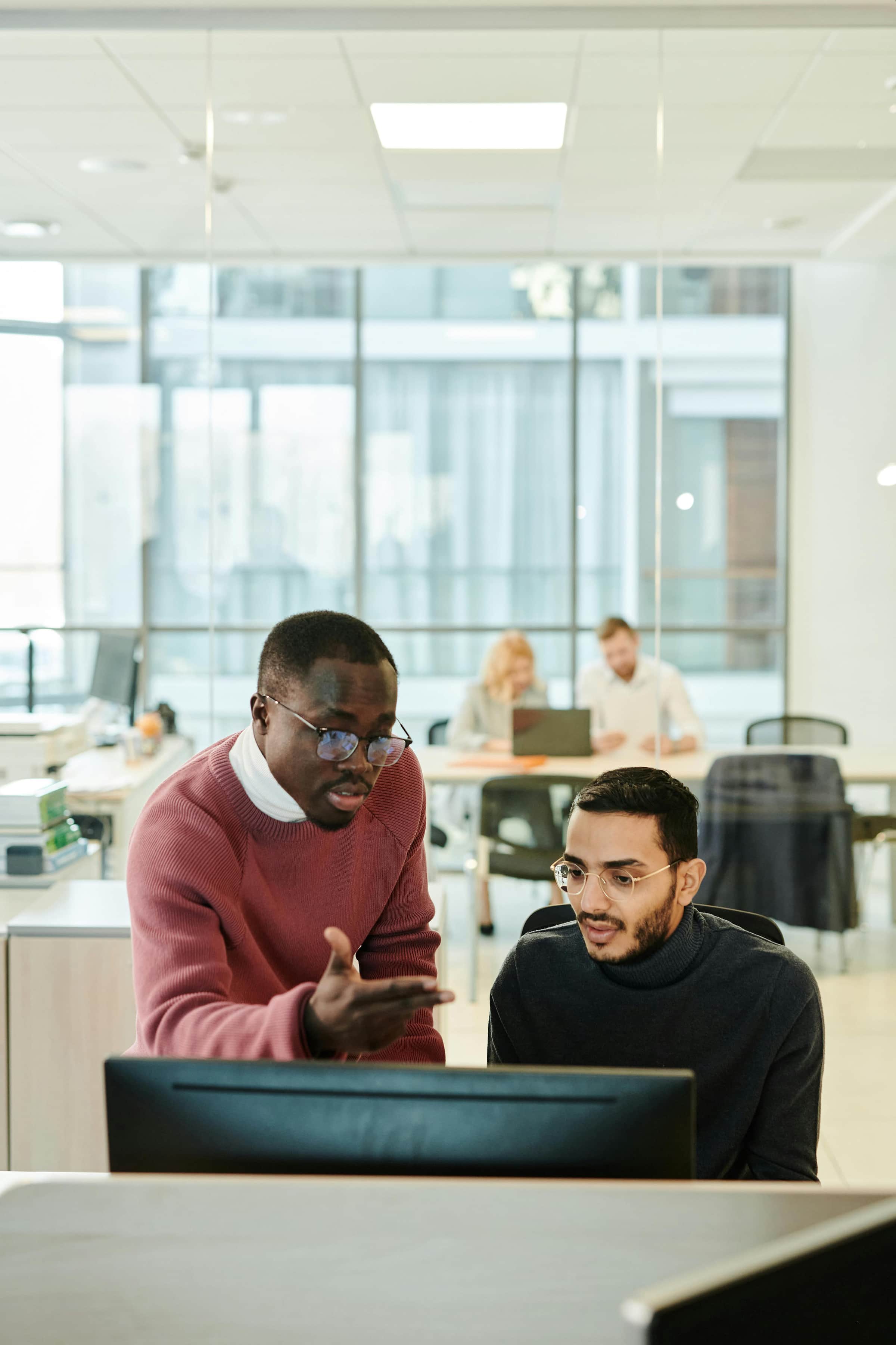 Foto de dois homens discutindo em frente a um computador em um escritório moderno, enquanto outros colegas trabalham ao fundo em um ambiente corporativo.