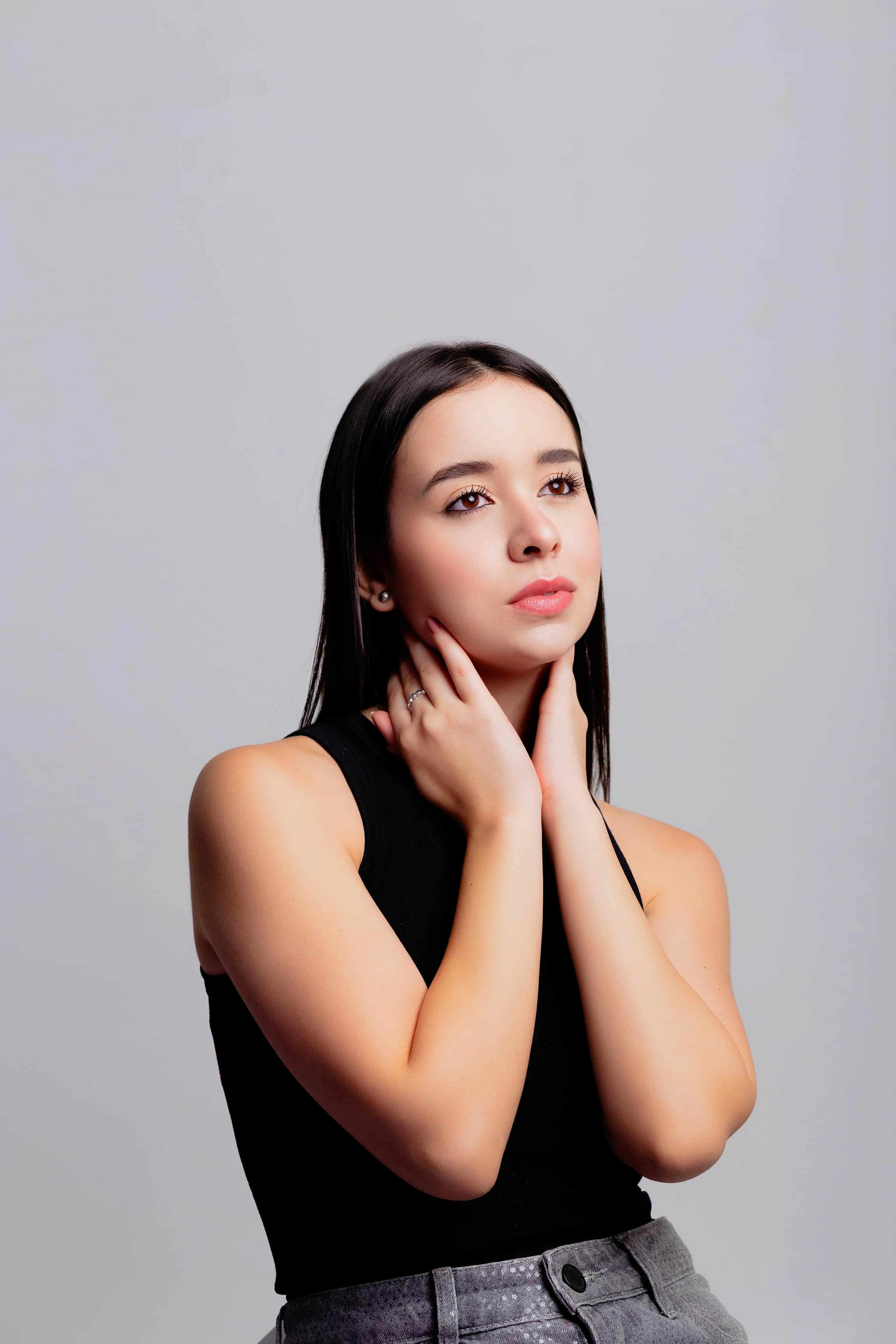 Foto de uma mulher jovem com pele clara e cabelos longos, posando elegantemente com as mãos no pescoço e usando roupa preta contra fundo neutro.