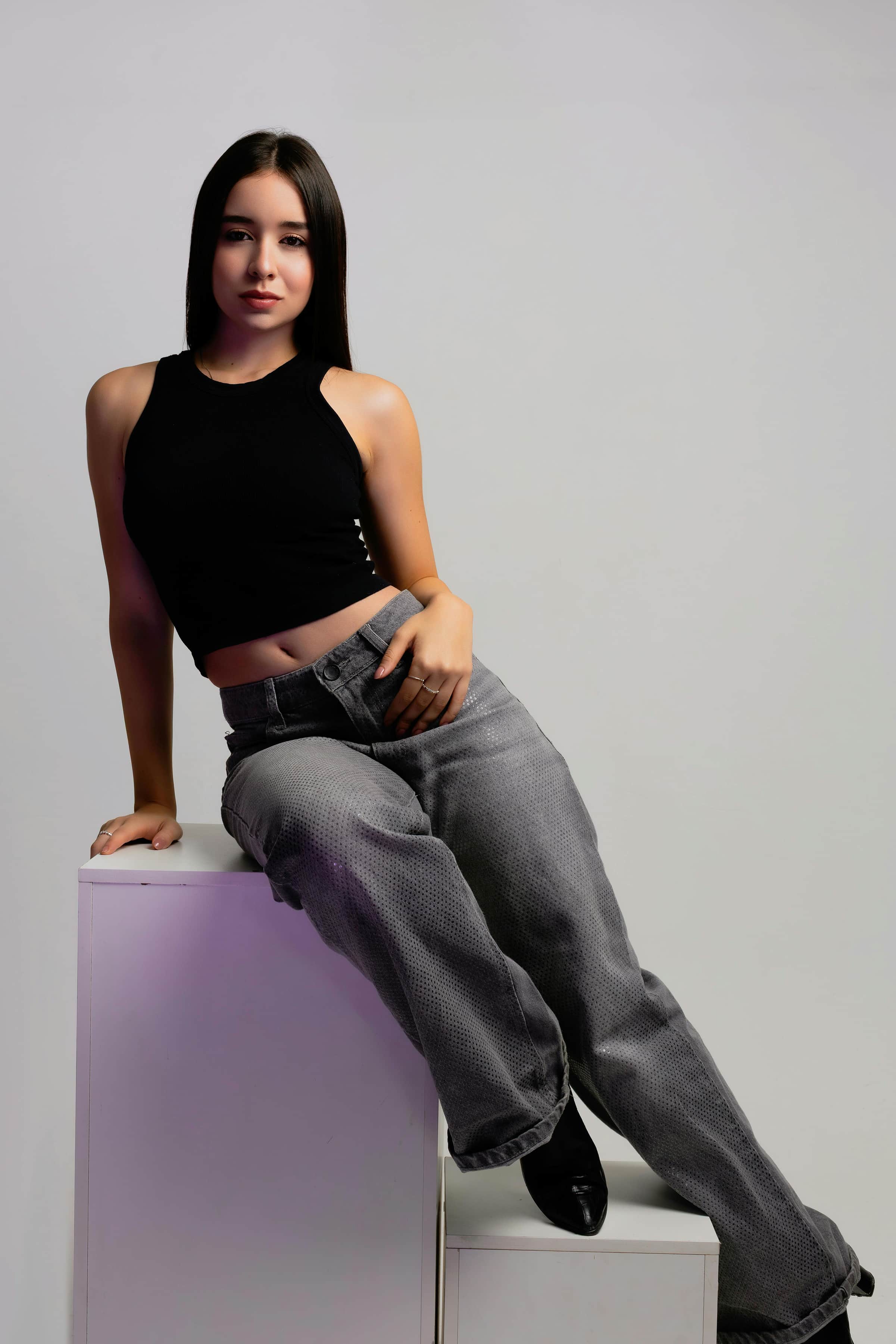 Foto de uma jovem mulher com pele clara e cabelos longos, vestindo uma blusa preta e calça larga e sentada de forma casual contra uma parede clara.