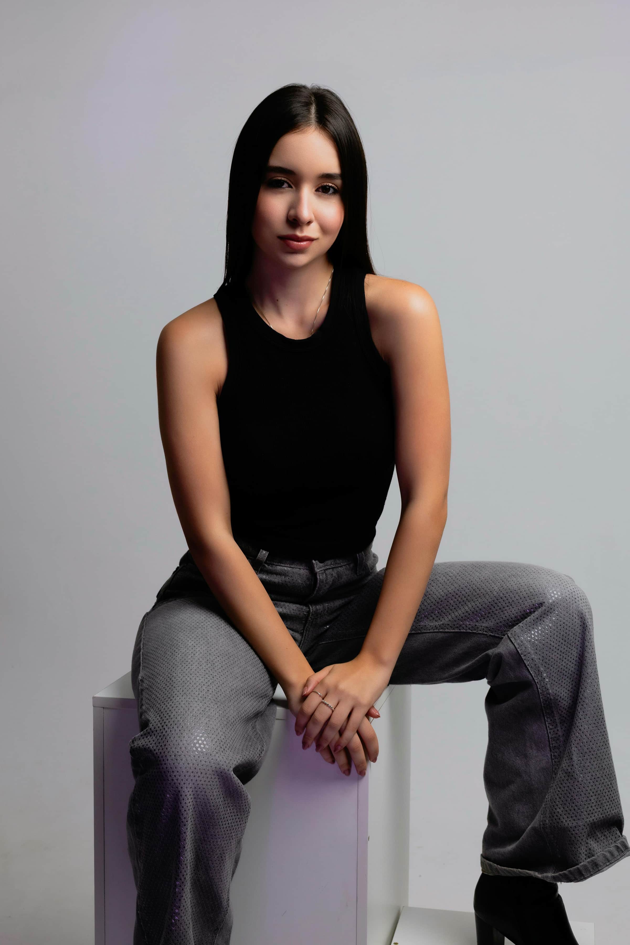 Foto de uma jovem mulher com pele clara e cabelos longos, sentada em uma posição casual e vestindo uma blusa preta e calças estilo jeans.