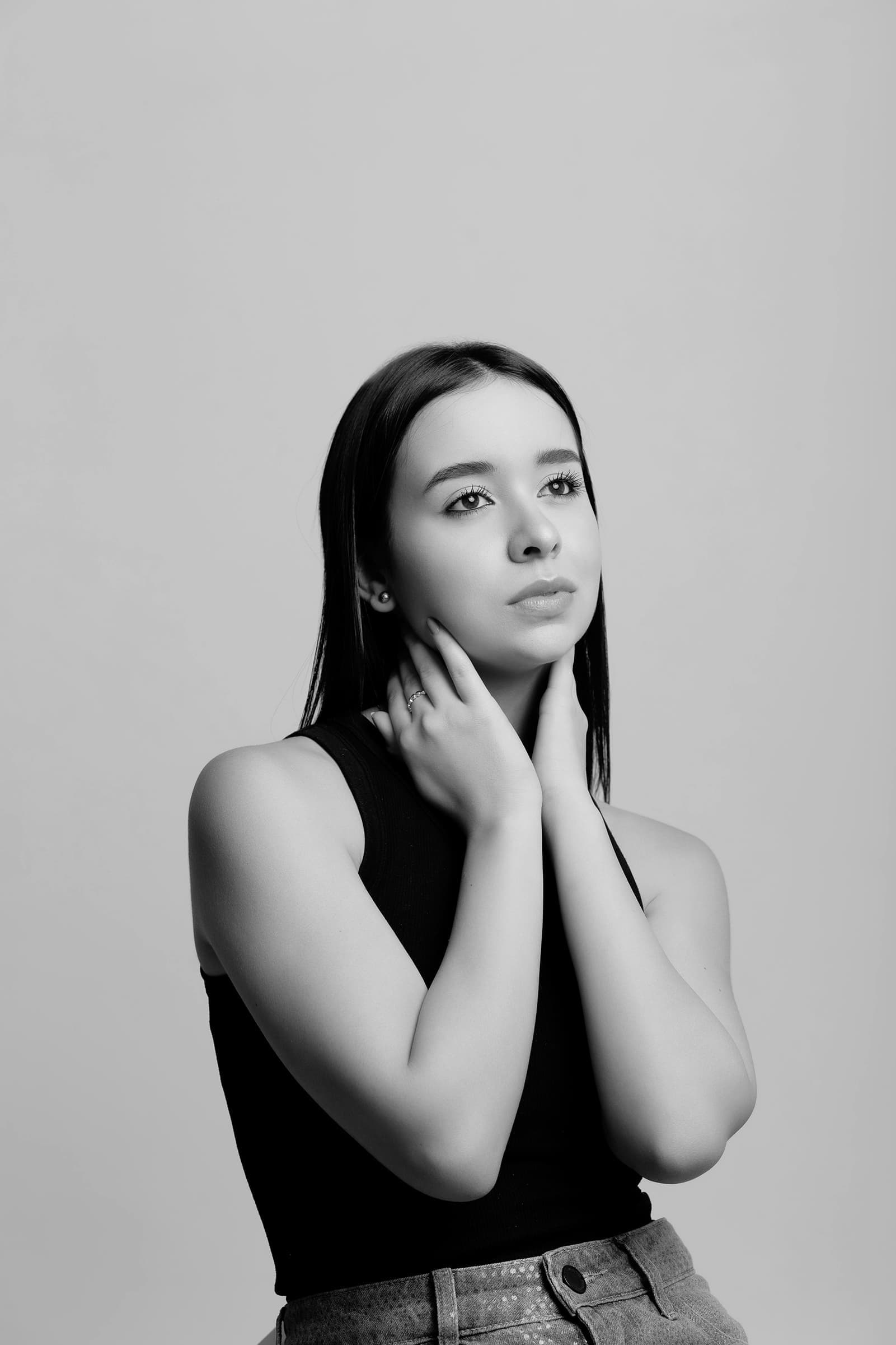 Foto de uma mulher jovem com pele clara e cabelos longos, posando elegantemente com as mãos no pescoço, usando roupa preta contra fundo neutro, em preto e branco.