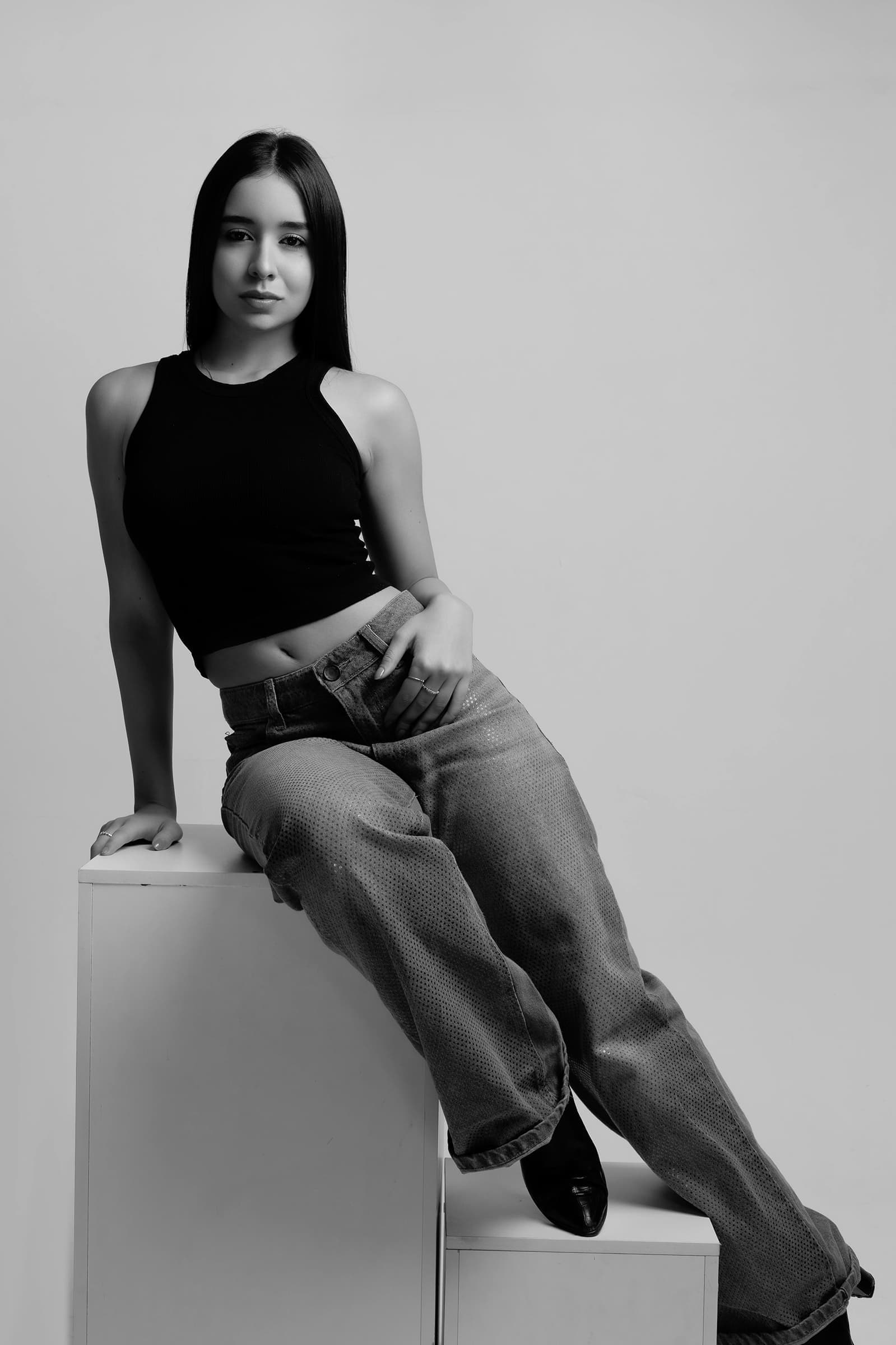 Foto de uma jovem mulher com pele clara e cabelos longos, vestindo uma blusa preta e calça larga, sentada de forma casual contra uma parede clara, em preto e branco.