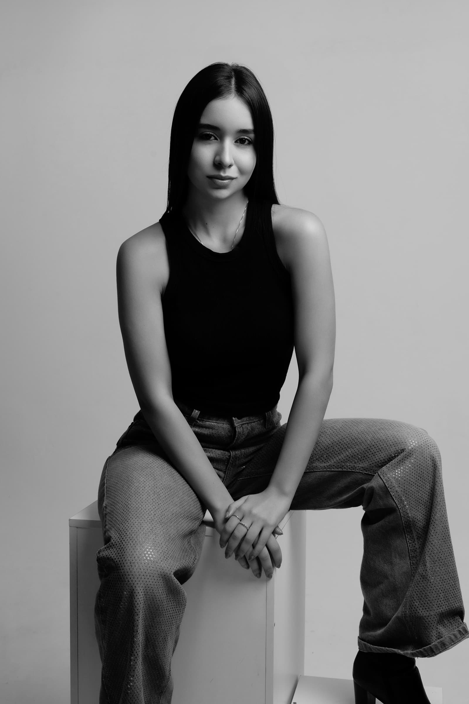 Foto de uma jovem mulher com pele clara e cabelos longos, sentada em uma posição casual, vestindo uma blusa preta e calças estilo jeans, em uma foto em preto e branco.