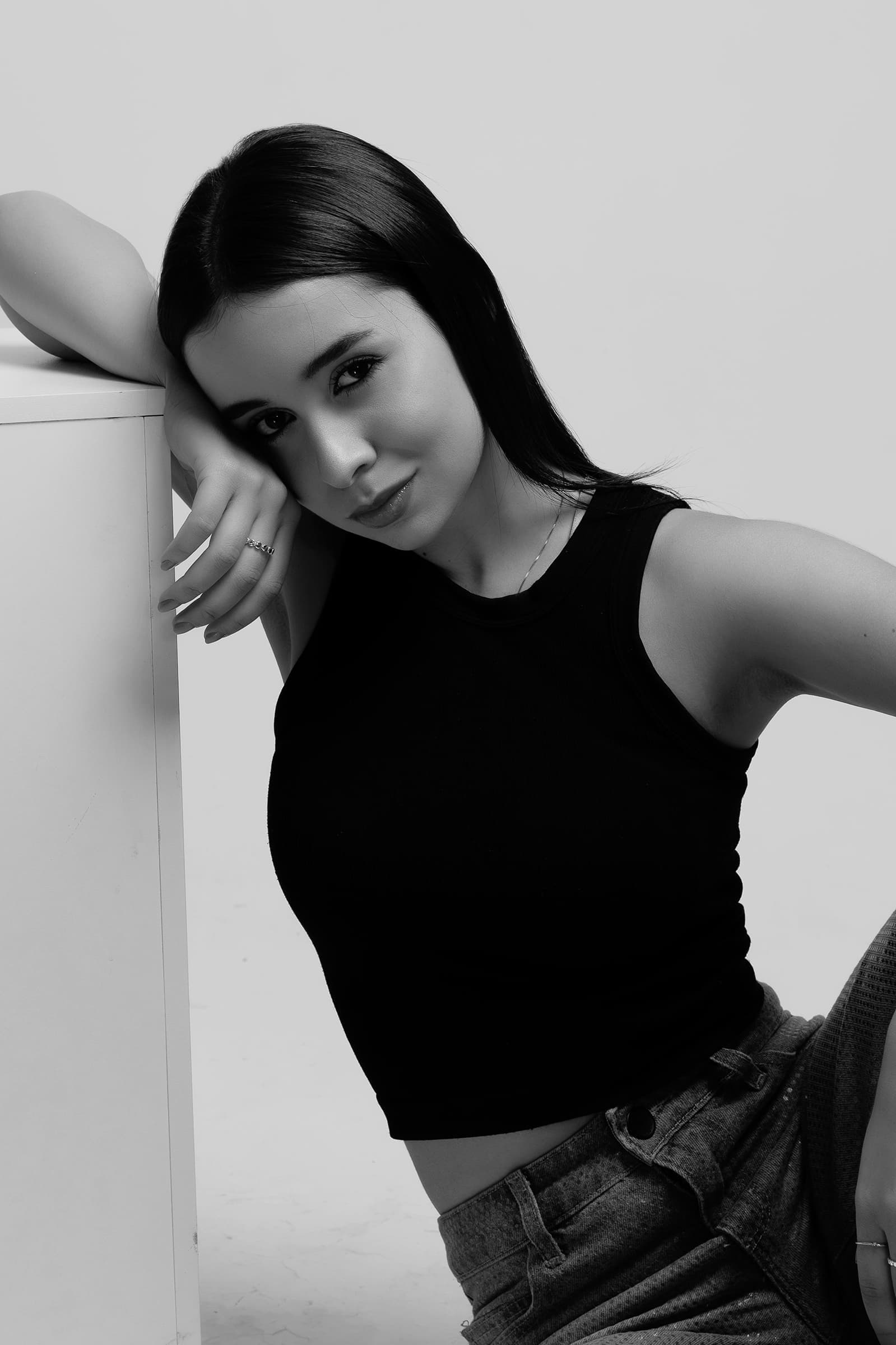 Foto de uma jovem mulher com pele clara e cabelos longos, usando top preto e jeans, posando de forma casual e moderna em preto e branco.