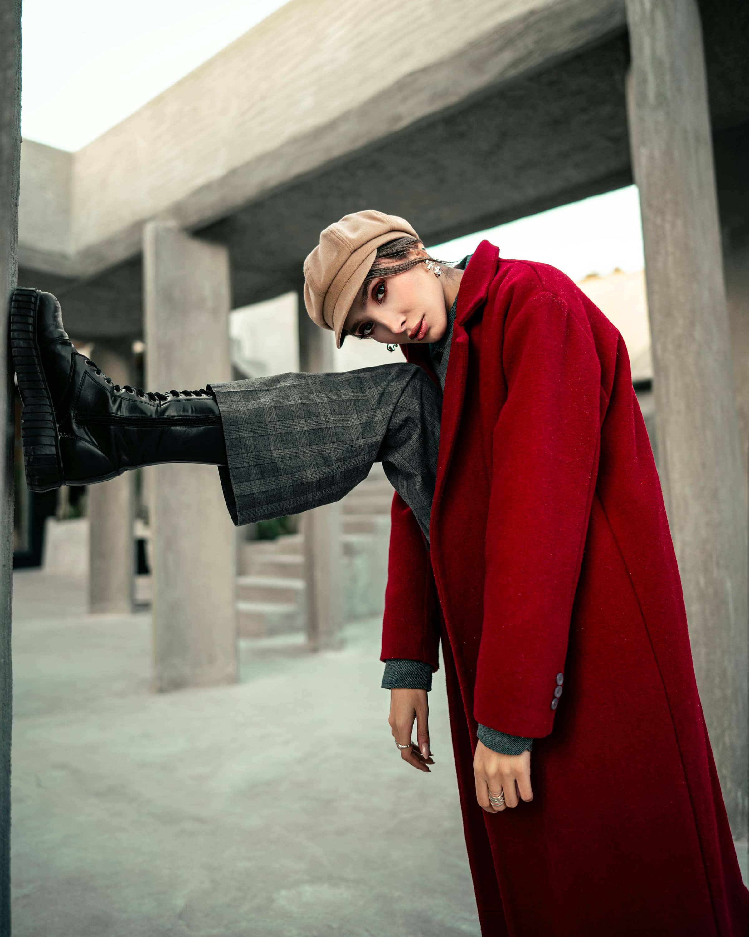 Foto de uma mulher estilosa com casaco vermelho, chapéu bege e calça de xadrez, posando de forma criativa sob estrutura de concreto moderna.