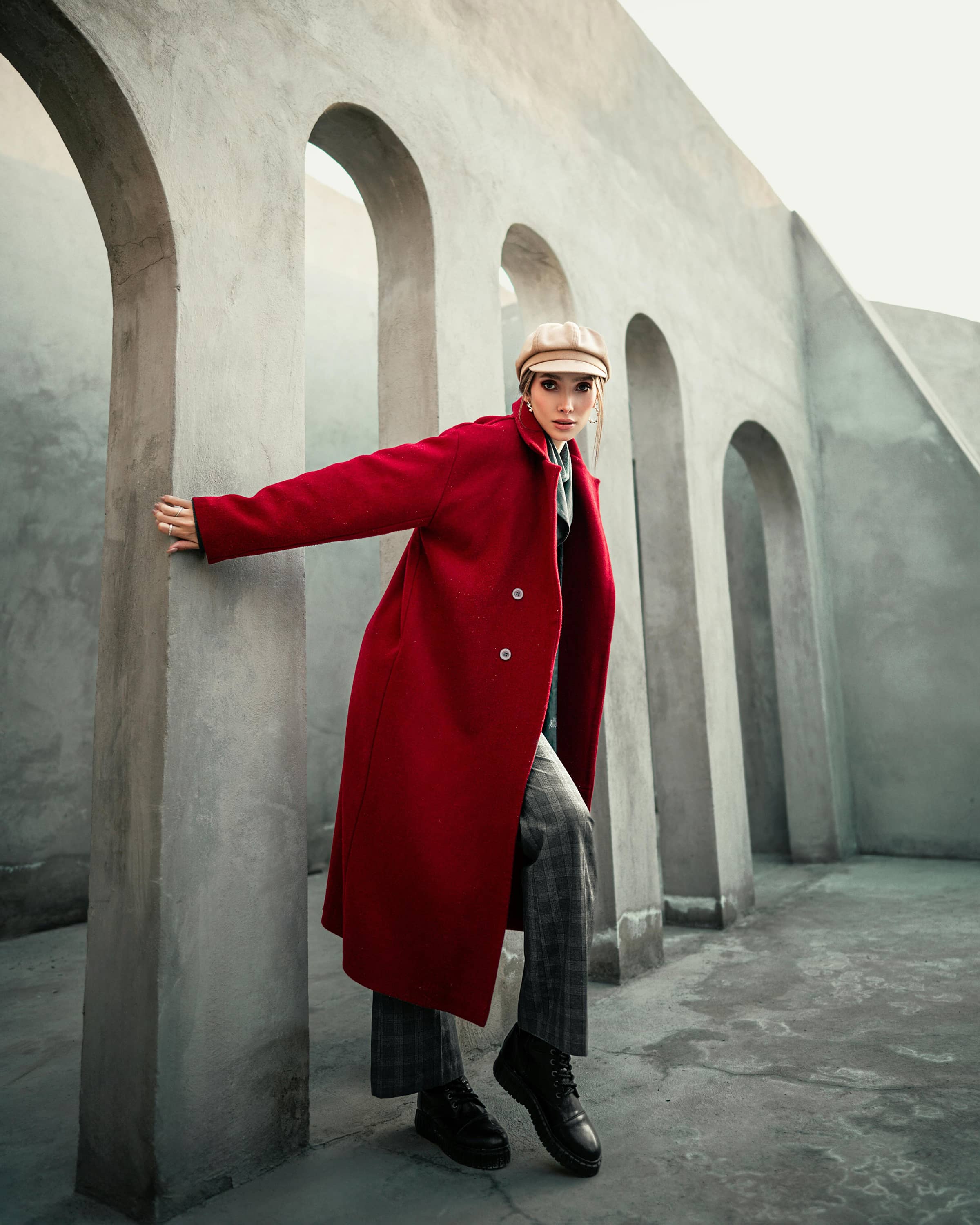 Foto de uma mulher usando casaco vermelho, calça xadrez e chapéu bege posando ao lado de paredes de concreto com arcos em cenário urbano moderno.