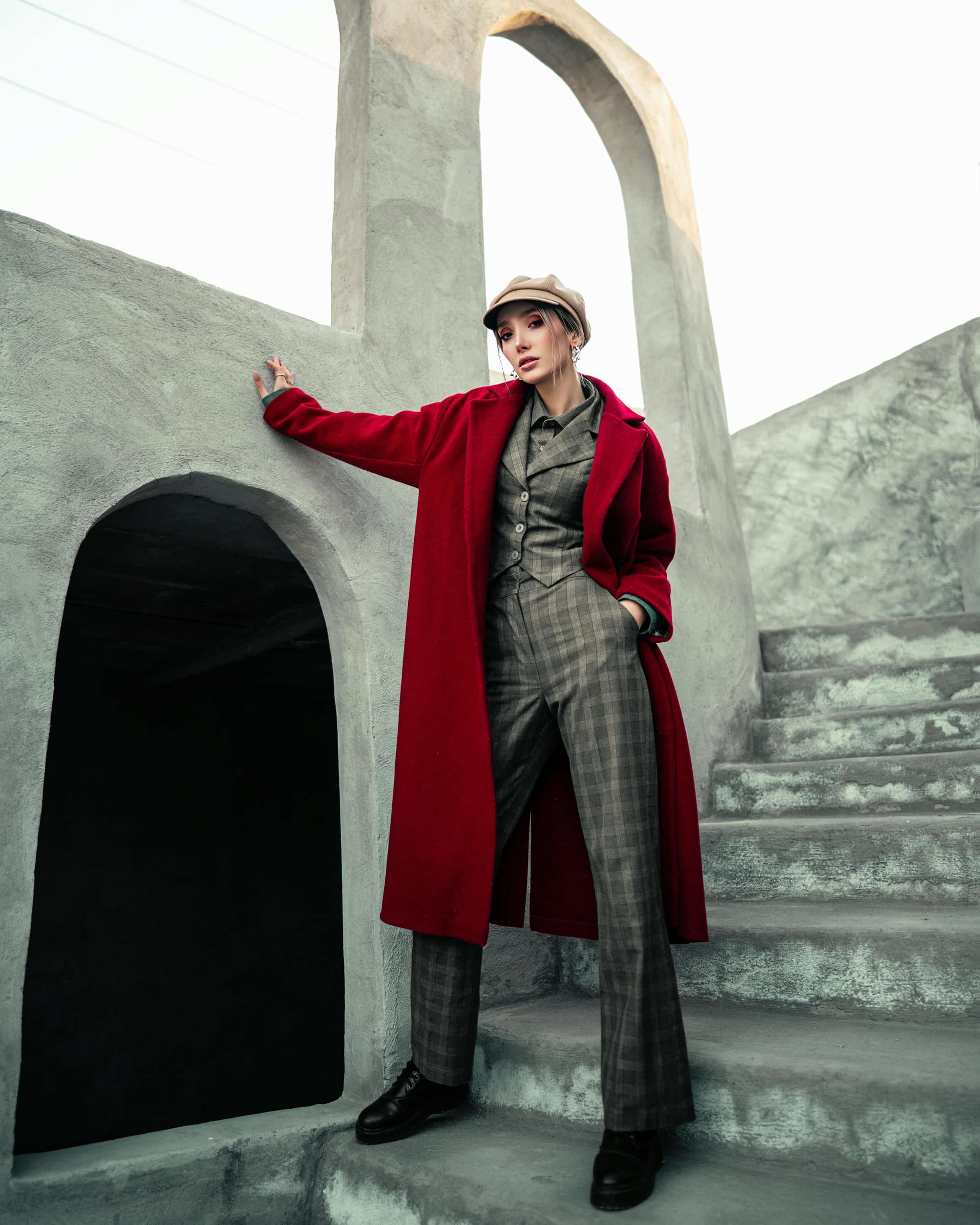 Foto de uma mulher usando casaco vermelho, chapéu bege e calça xadrez posando em escadaria de concreto em cenário urbano