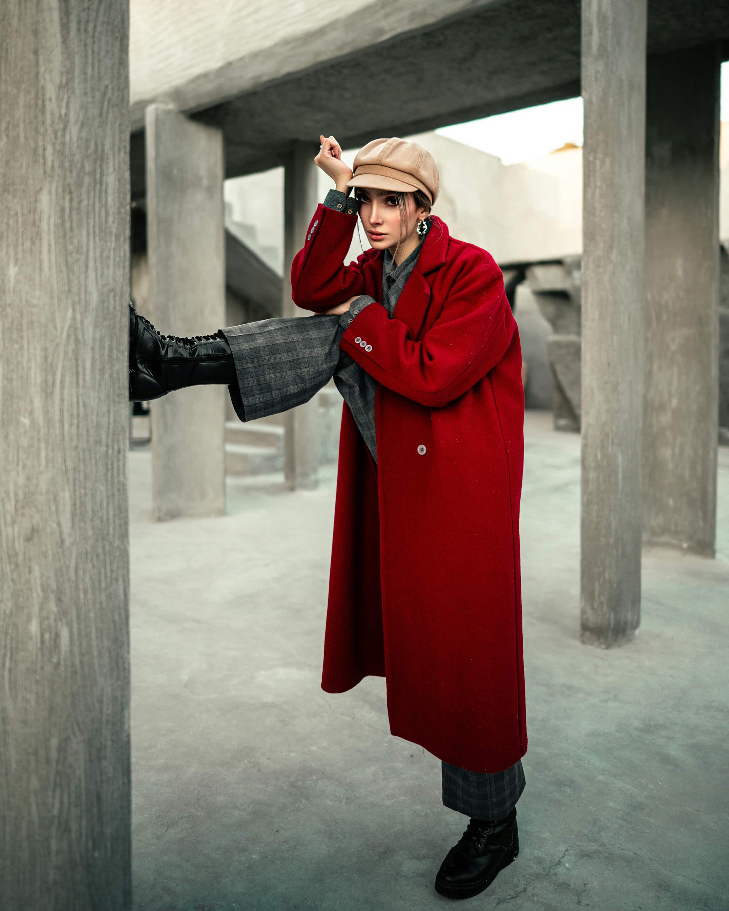 Foto de uma mulher com estilo fashion usando blazer vermelho longo, calça xadrez, bota preta e chapéu bege em ambiente urbano com estrutura de concreto
