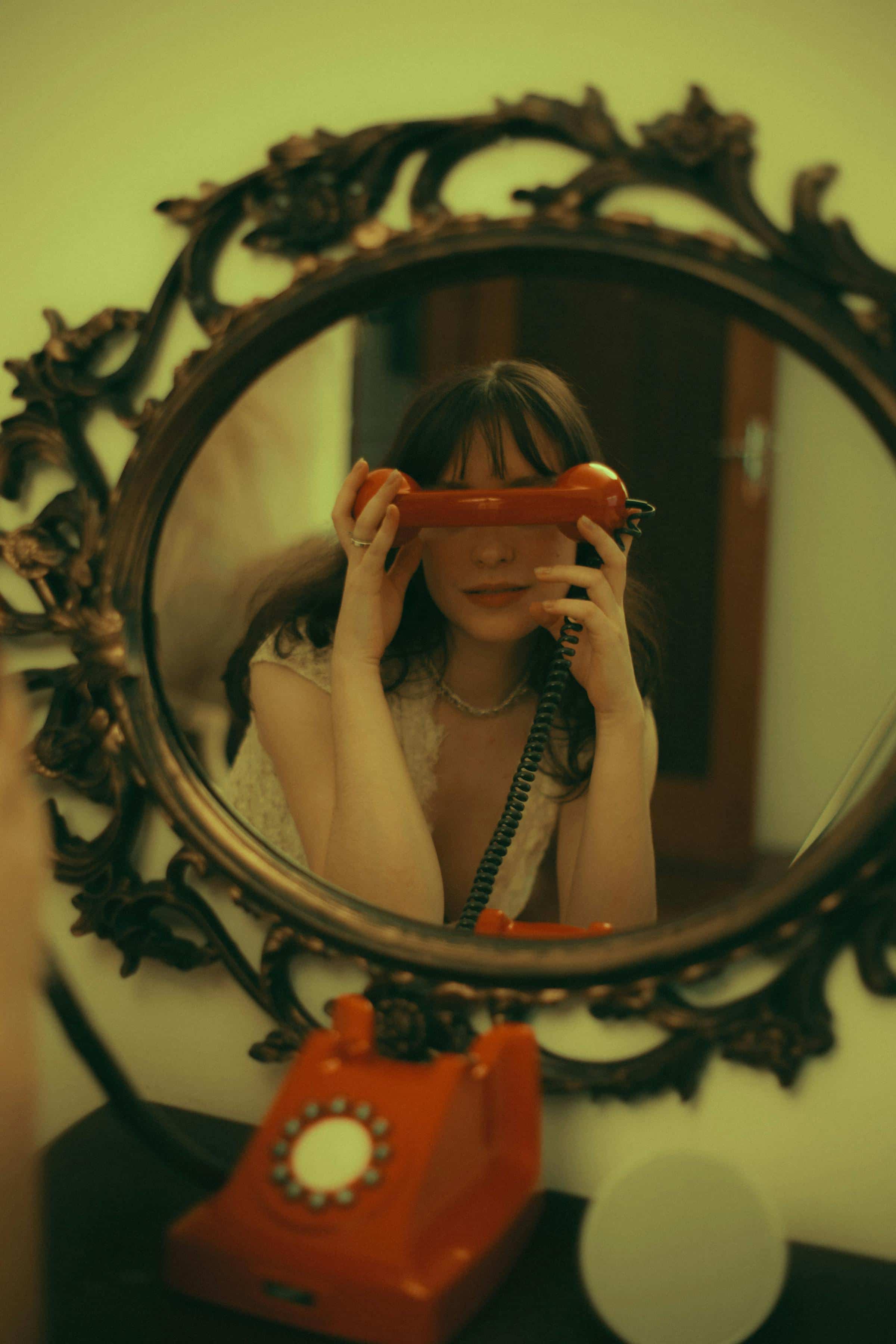 Foto de uma mulher com cabelo escuro segurando telefone antigo em um espelho decorado, com iluminação quente, estilo vintage.