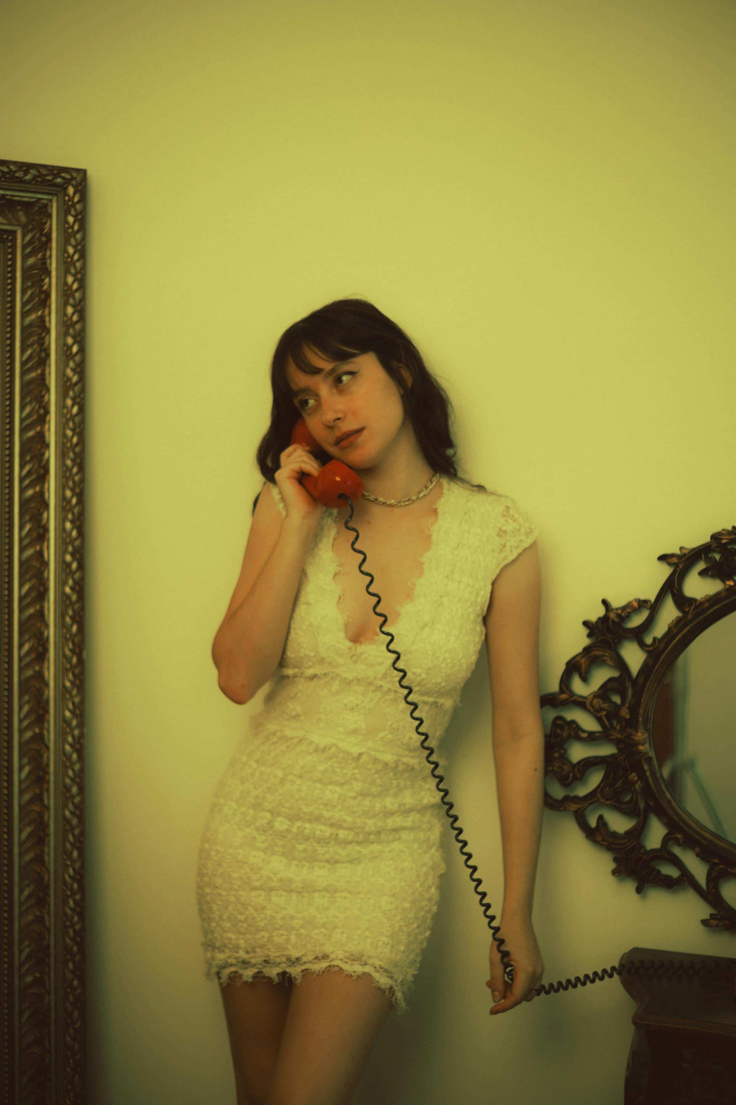 Foto de uma mulher com cabelo escuro e vestido branco, segurando telefone antigo vermelho na orelha, ao lado de espelho decorativo na parede.