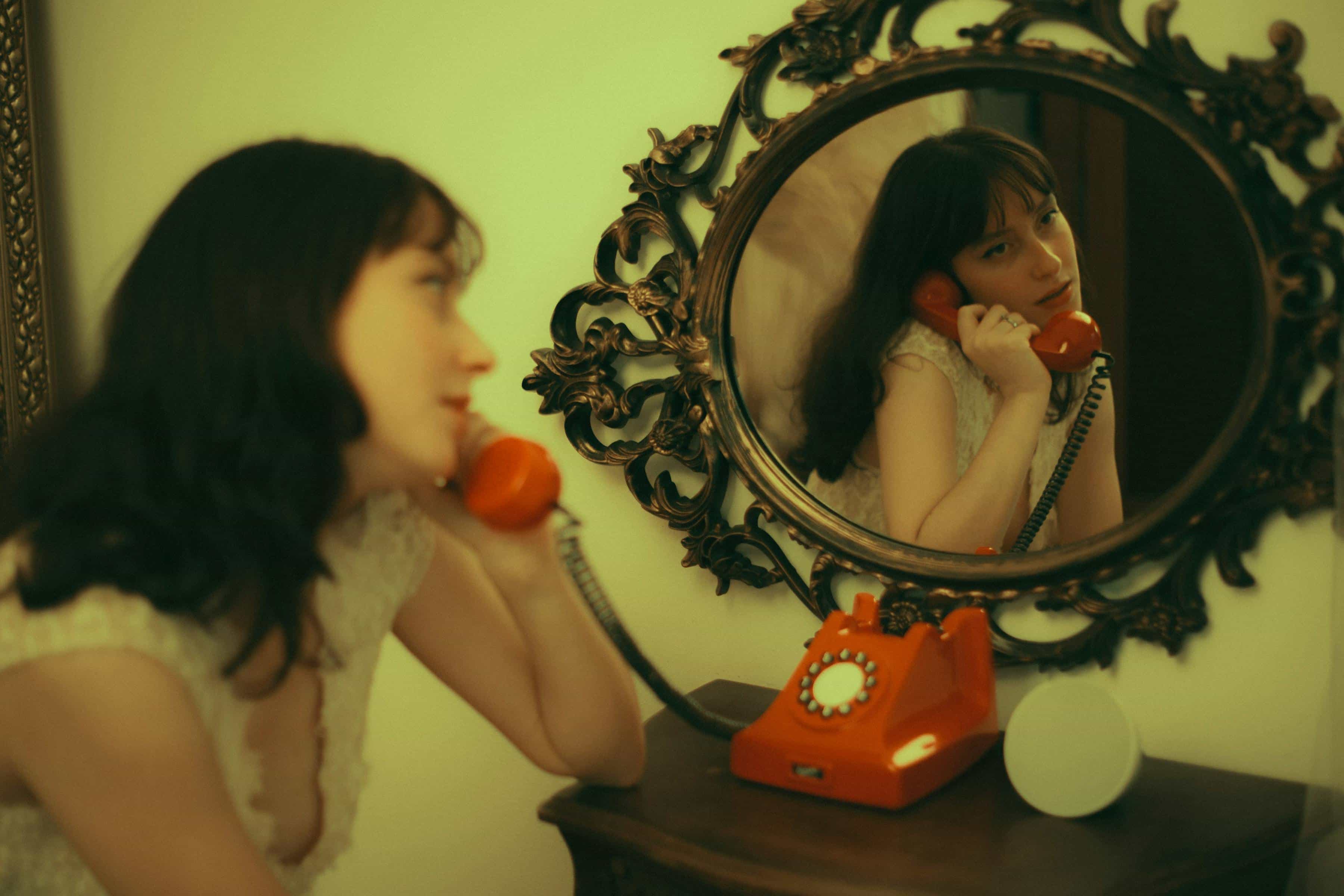 Foto de uma mulher usando blusa branca falando ao telefone antigo vermelho enquanto olha no espelho, refletindo seu rosto.