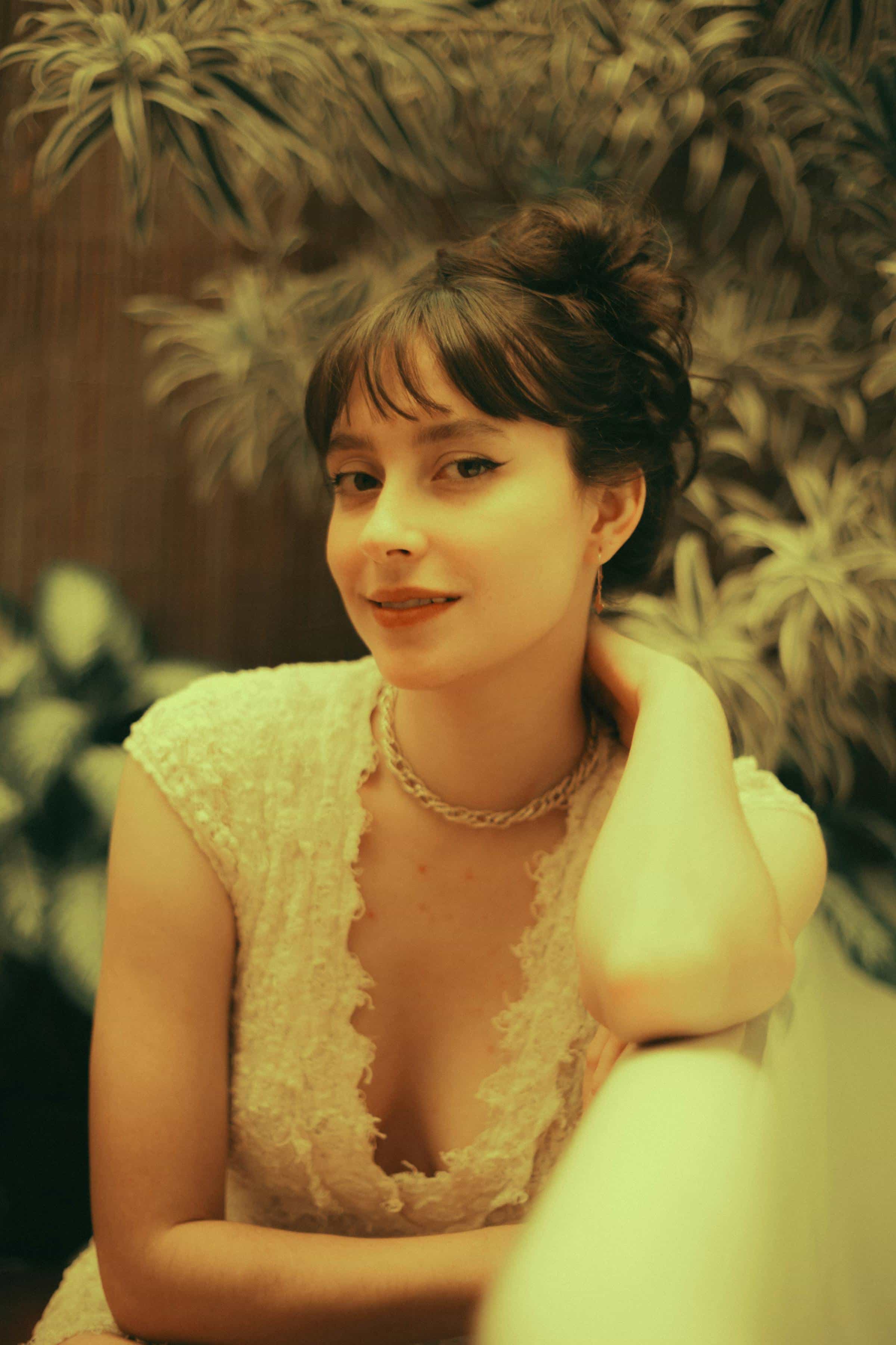 Foto de uma mulher com cabelo escuro preso em um coque bagunçado, sorrindo suavemente, usando vestido de renda branco, posando diante de um fundo com plantas verdes, com iluminação quente, estilo vintage.
