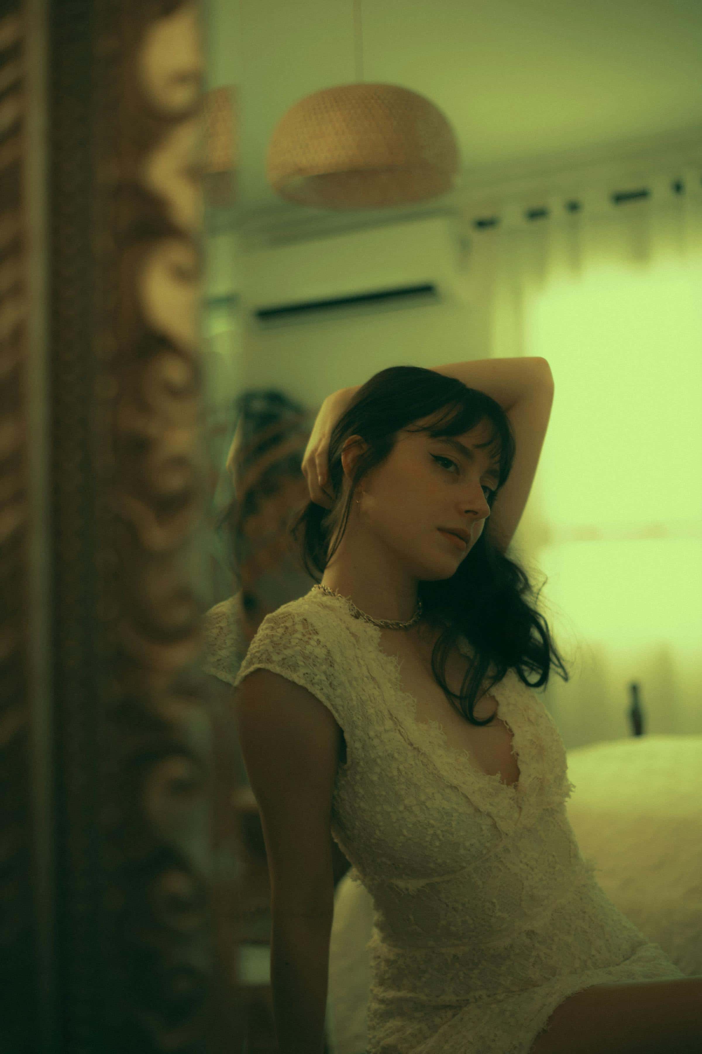 Foto de uma mulher com cabelo escuro e vestido de renda branco posando em ambiente com iluminação amarelada, refletindo no espelho e criando uma atmosfera íntima.