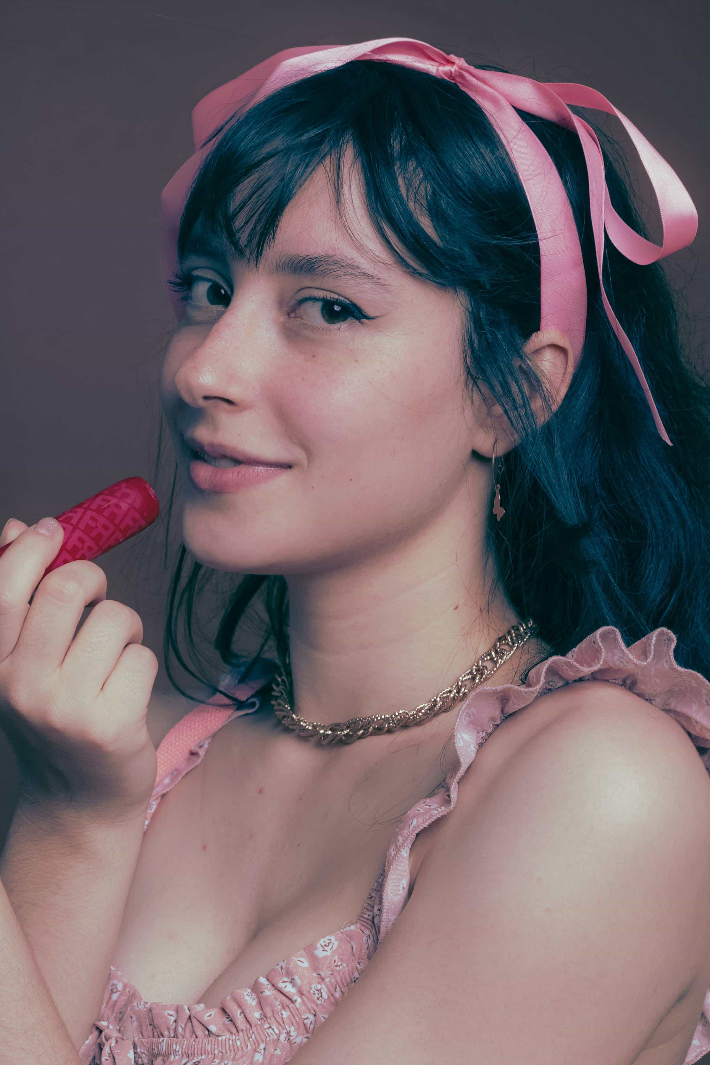 Foto de uma mulher com cabelo escuro usando faixa rosa no cabelo, vestindo roupa rosa, segurando um batom, com um leve sorriso.