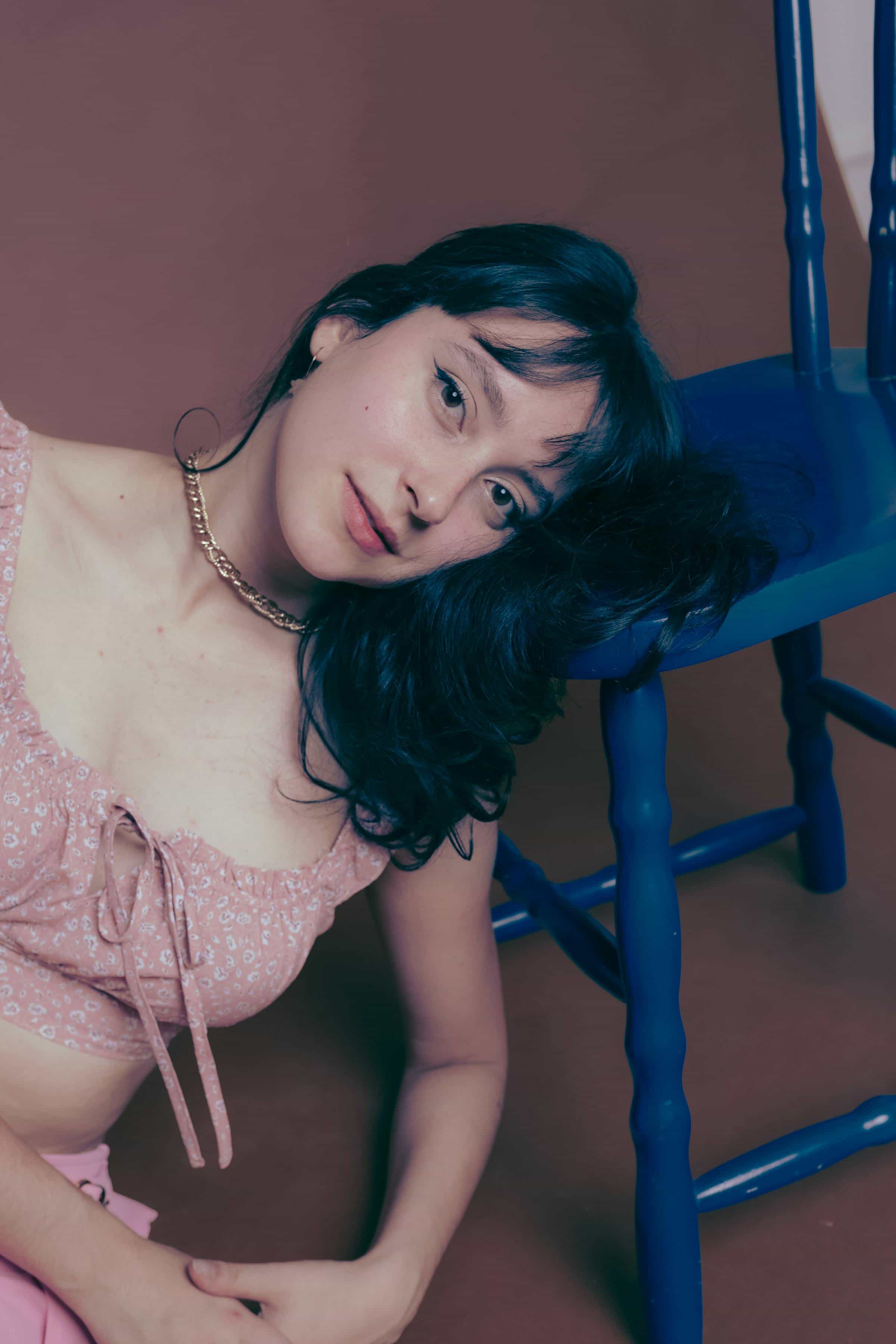 Foto de uma mulher com cabelo escuro usando blusa rosa com detalhes florais e acessórios dourados, posando próxima a uma cadeira azul.