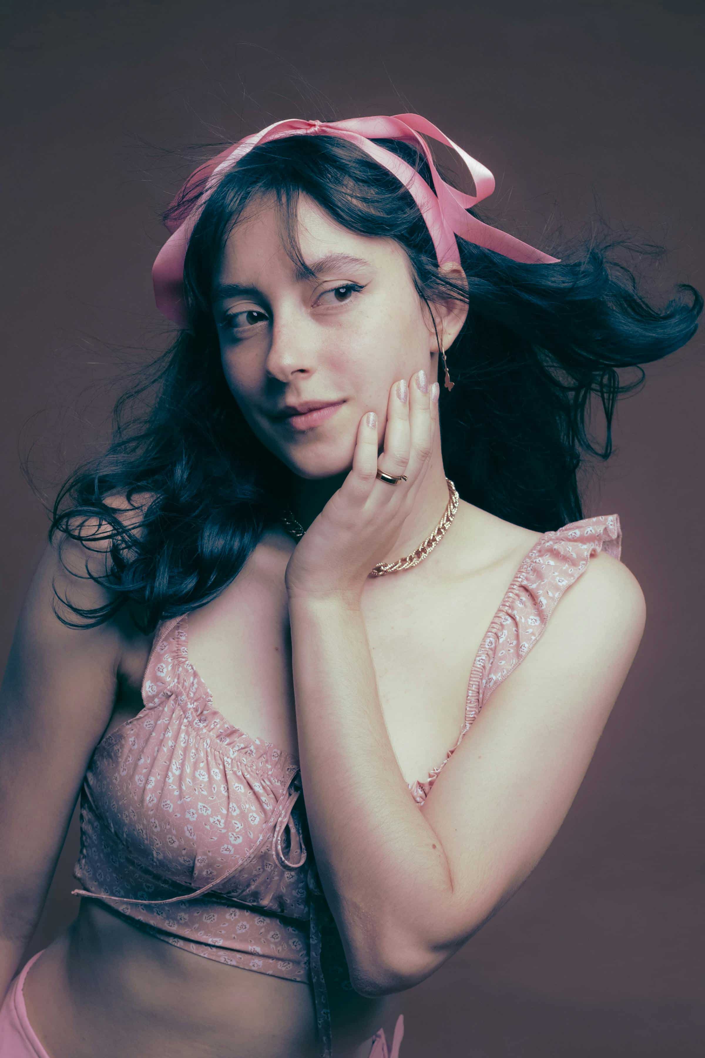 Foto de uma mulher com cabelo escuro, usando acessórios dourados, um vestido rosa com detalhes em renda, e um laço rosa na cabeça.