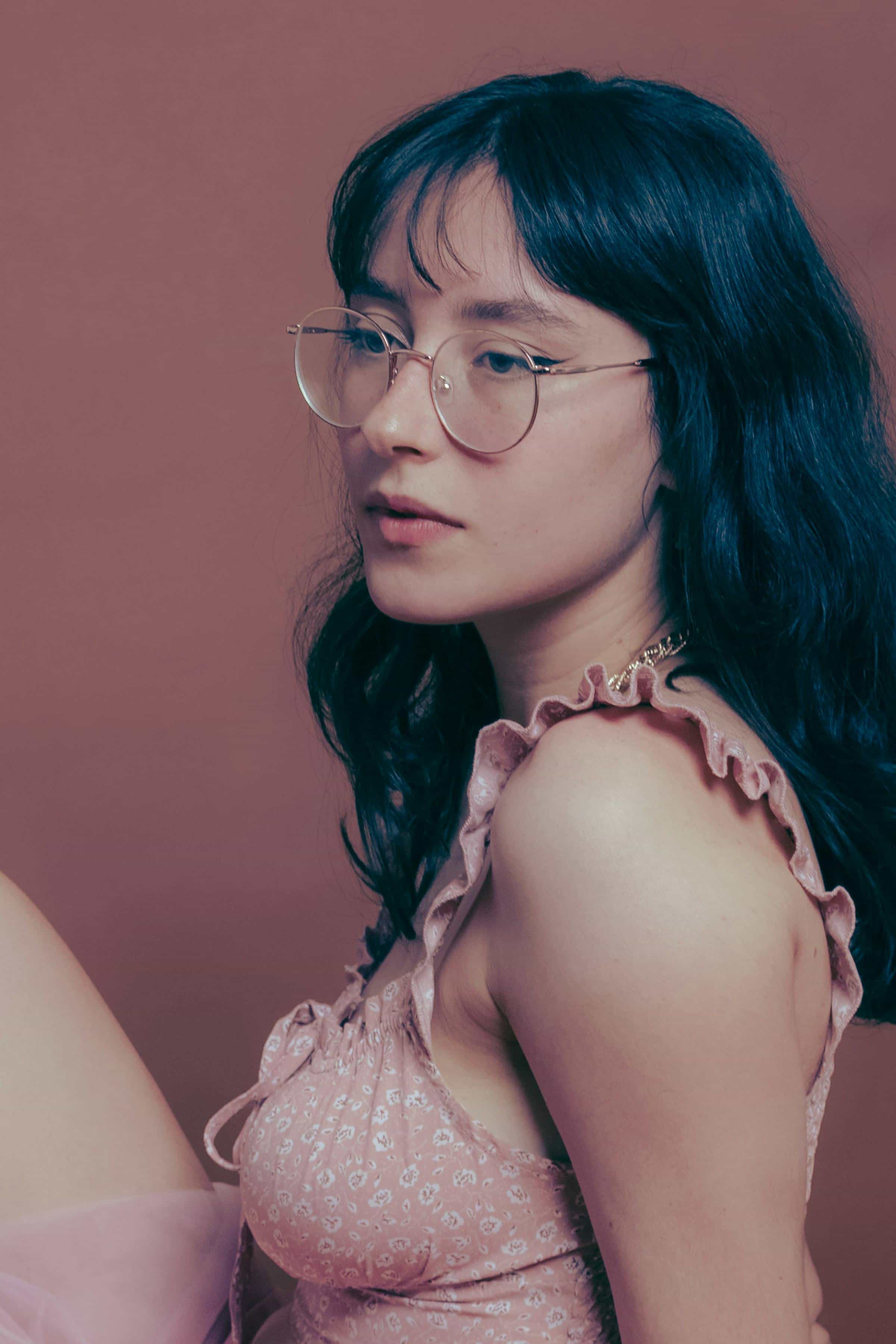 Foto de uma mulher jovem com cabelo escuro, usando óculos, vestido rosa e colar de corrente.