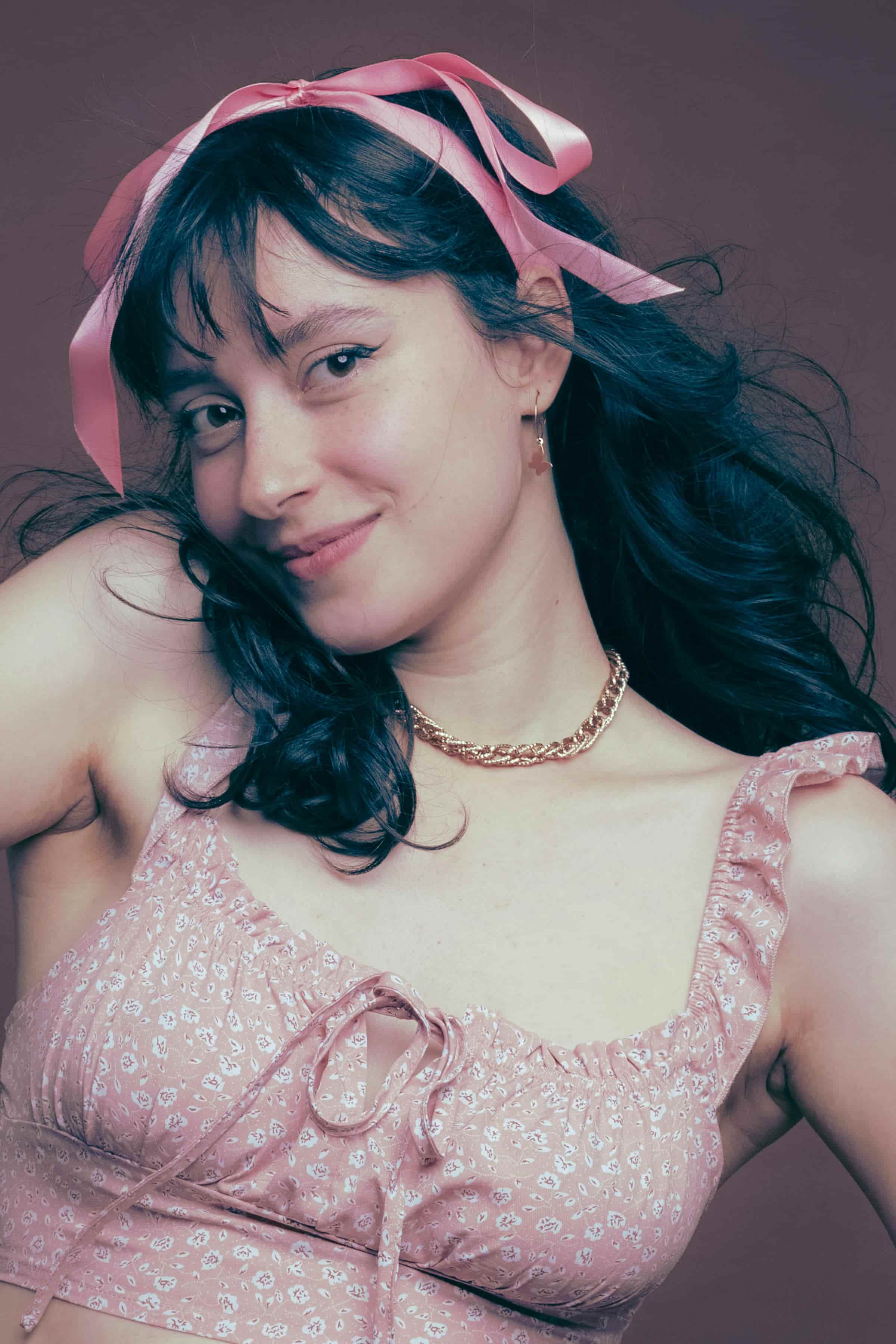 Foto de uma mulher jovem com cabelo escuro, usando laço rosa na cabeça, vestido rosa e colar de corrente, sorrindo para a câmera.