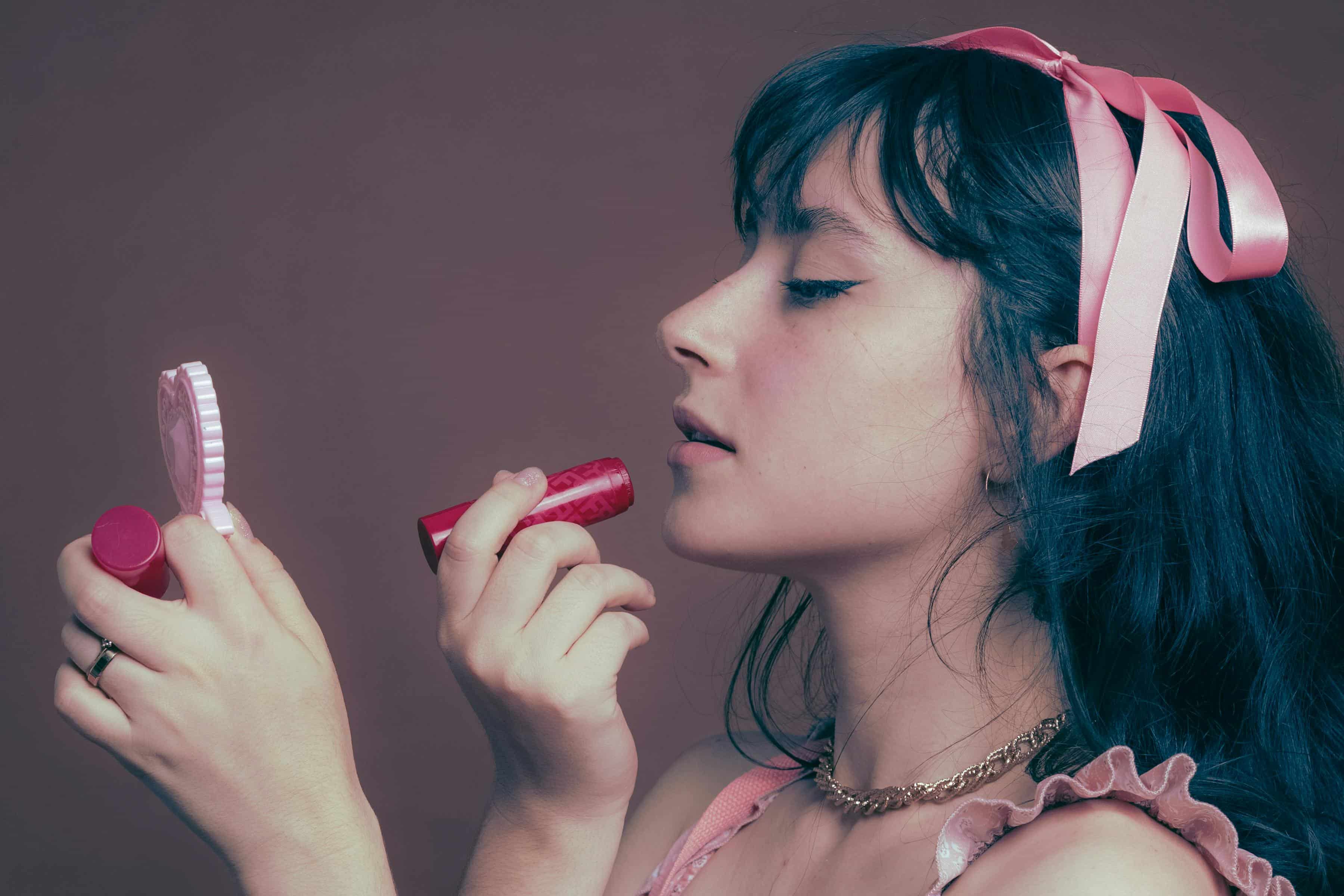 Foto de uma mulher com cabelo escuro, usando laço rosa na cabeça, se maquiando, com foco na maquiagem e beleza.